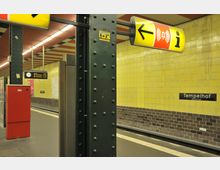 U-Bahn-Station mit gelben Kacheln an den Wänden, schwarzen Säulen mit Nieten, einem rot-orangefarbenen Alarmkasten und Richtungsschildern. Im Hintergrund ist das Stationsschild "Tempelhof (Südring)" und die Bahnsteigkante zu sehen.