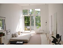 Helles Zimmer mit weißen Wänden und hohen Glasfenstern, die auf einen Balkon mit Topfpflanzen führen. Es gibt eine niedrige Matratze mit hellen Kissen auf dem Boden und minimalistische Möbel, darunter ein Spiegel an der Wand und einige Pflanzen.
