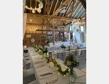 Rustikale Veranstaltungshalle mit Holzbalkendecke, dekoriert für eine Hochzeit oder Feier. Lange Tische mit weißen Tischdecken, Blumengestecken und Lichterketten; im Hintergrund eine Bar mit Backsteinwand und Fenstern mit Tageslicht.