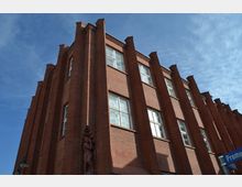 Backsteinfassade eines mehrstöckigen Gebäudes im Stil des Expressionismus mit vertikalen Ziegelsäulen und weißen Fenstern, darunter eine kleine Skulptur an der Ecke. Der Himmel ist klar und blau, ein Straßenschild mit der Aufschrift „Promenade“ ist unten rechts sichtbar.