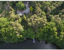 Luftaufnahme eines Hauses mitten in einem Wald mit dichtem grünen Baumkronen, in der Nähe eines Fluss- oder Seeufers. Ein Boot liegt am Wasser und ein Steg ist sichtbar, umgeben von natürlicher Vegetation.