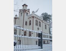 Schnee bedeckte Fassade eines historischen Gebäudes im neugotischen Stil mit weißen Wänden und rotbraunen Backsteinverzierungen; im Vordergrund ein schmiedeeiserner Zaun, ebenfalls mit Schnee bedeckt, und am oberen Bildrand winterlich kahle Bäume.