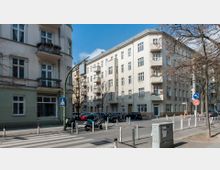 Straßenecke in einem Wohngebiet mit mehrstöckigen Altbauhäusern, Balkonen und stehenden Autos sowie Motorrollern vor den Gebäuden. Im Vordergrund befinden sich ein Zebrastreifen, Verkehrszeichen und vereinzelt stehende Bäume.