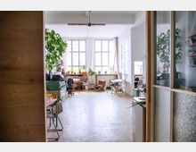 Heller Atelierraum mit großen Fenstern, hohen Decken und einer Einrichtung aus Schreibtischen, Regalen, Bürostühlen sowie Pflanzen. Im Vordergrund sind Holztüren und Glaswände sichtbar, die den Raum abtrennen.