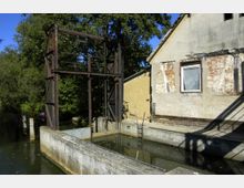 Eine alte Schleusenanlage mit verwitterten Betonwänden und einer Metallkonstruktion im Vordergrund. Rechts daneben steht ein verfallenes Gebäude mit teils freiliegendem Mauerwerk, im Hintergrund sind Bäume sichtbar.