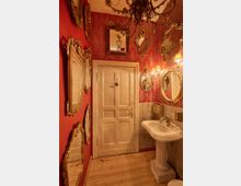 Kleines Badezimmer mit roten Wänden und goldverzierten Spiegeln, die in verschiedenen Formen an der Wand hängen. Der Raum wird von einem Kronleuchter und zwei Wandlampen erleuchtet und enthält ein weißes Waschbecken vor einer cremefarbenen Tür.