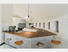 Moderne Küche in einem Gebäude mit Rundfenstern, einer geschwungenen Wand und einer hohen, weißen Decke. Die Einrichtung umfasst eine Arbeitsfläche mit Holzplatte, Barhocker sowie eine eingebaute Spüle und Kaffeemaschine.