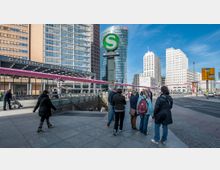 Ansicht des Potsdamer Platzes in Berlin mit modernen Hochhäusern im Hintergrund, dem Eingang zur S-Bahn-Station "Potsdamer Platz" im Vordergrund und mehreren Fußgängern auf dem Platz. Eine markante pinkfarbene Rohrleitung verläuft quer durch das Bild.