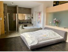 Schlafzimmer mit integriertem Badezimmer, bestehend aus einer grauen Polsterliege mit bedruckter Bettdecke im Vordergrund, einer freistehenden Badewanne, einem Waschtisch mit Spiegel sowie einer verglasten Dusche im Hintergrund. Links sind Ziegelwände und rechts ein Regal mit Blumen und Lichtakzenten sichtbar.