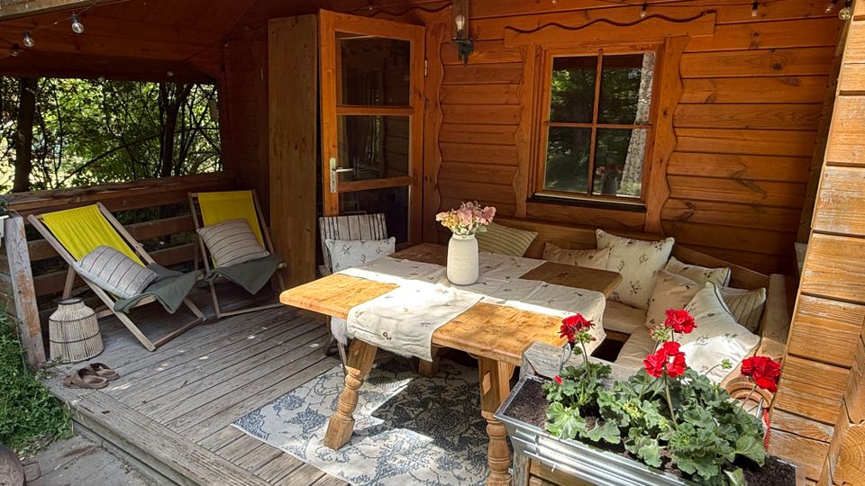 Überdachte Holzterrasse einer Blockhütte mit Holzdecke und Lichterkette, offener Tür und Fenster. Auf der Veranda stehen zwei Liegestühle, eine Eckbank mit Kissen und ein Holztisch mit Tischläufer und Vase; vorn ein Blumenkasten mit roten Geranien.