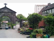 Historische Steinmauer mit Torbogen und Kreuz darüber, flankiert von üppiger Begrünung. Im Vordergrund stehen farbenfrohe Blumenkübel und Pflanzen, links ein Eisengitter, rechts ein Gebäude mit rotem Putz und Ziegeldach.