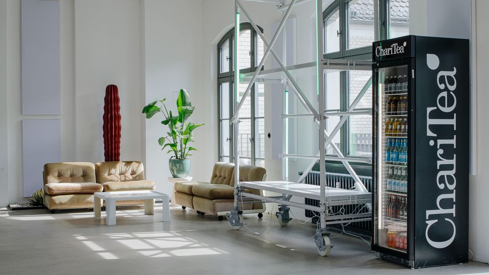 Heller, minimalistischer Loft-Raum mit hohen Decken, großen Rundbogenfenstern und glattem Betonboden. In einer Sitzecke stehen beigefarbene Sofas, ein niedriger Tisch und Pflanzen; daneben ein fahrbares Gerüst und ein Getränkeautomat an der Wand.