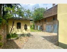 Kleiner Innenhof mit gepflastertem Boden, umgeben von gelb gestrichenen und unverputzten Backsteingebäuden mit blauen Holztüren. Ein Baum wächst neben einem gelben Gebäude mit einem kleinen Fenster, in dem rote Blumen stehen.