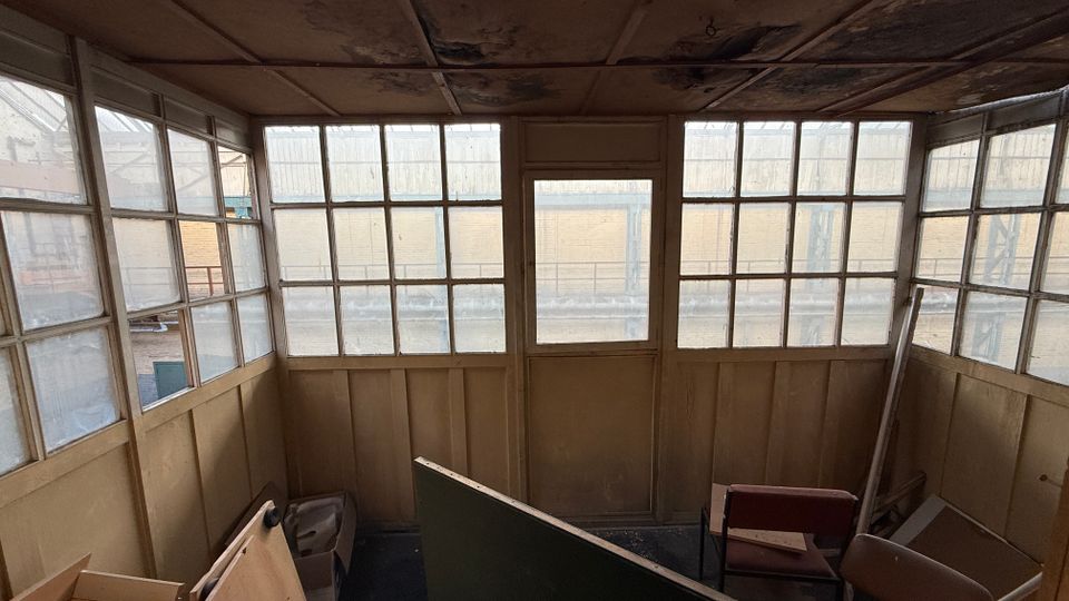 Innenraum einer verwitterten, verglasten Hütte oder eines Warteraums mit großen, in Raster unterteilten Fenstern an drei Seiten und einer Tür in der Mitte. Die Decke ist fleckig und beschädigt, im Raum stehen verstreute Möbelteile und Bretter.