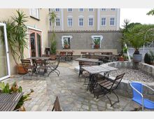 Ein gepflasterter Außenbereich mit mehreren Holz-Tischen und Stühlen auf Metallgestellen, umgeben von Topfpflanzen, darunter Palmen und andere Grünpflanzen. Im Hintergrund ist eine helle Gebäudefassade mit mehreren Fenstern zu sehen.