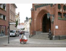 Straßenszene in Alt-Köpenick mit einem roten Backsteingebäude mit gotischem Spitzbogenportal auf der rechten Seite, flankiert von einer Laterne und einer Statue. Im Hintergrund sind eine gepflasterte Straße, parkende Autos und Wohnhäuser zu sehen.