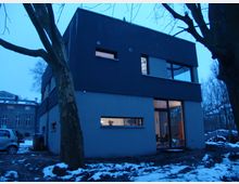 Modernes zweistöckiges Wohnhaus mit dunkler Fassade und großen Fenstern, umgeben von verschneitem Boden und Bäumen in einer städtischen Umgebung. Im Hintergrund sind weitere Gebäude und parkende Autos zu sehen.