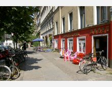 Straßenszene in einer urbanen Umgebung mit mehrstöckigen Altbauten; auf der rechten Seite ein Geschäft mit roter Fassade ("Doppelglück Impex") und ausgestellten Waren wie bunten Stühlen. Links sind Fahrräder geparkt, daneben ein Gehweg mit Bäumen, Tischen und Personen unter einem blauen Sonnenschirm.