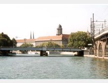 Blick auf einen Fluss mit einer Brücke im Vordergrund, umgeben von Gebäuden, darunter eine historische Struktur mit rotem Dach und einer Kuppel. Rechts neben der Brücke verlaufen Bahngleise auf erhöhten Bögen; im Hintergrund ragen Kirchtürme und Bäume hervor.
