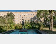 Eine Skulptur eines stehenden nackten Menschen auf einem Sockel in einem leeren Brunnen, umgeben von dichtem Grün und Bäumen. Im Hintergrund ist ein beige Wohngebäude mit mehreren Fenstern zu sehen.