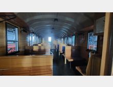 Innenansicht eines historischen Eisenbahnwaggons mit holzverkleideten Wänden, Sitzbänken aus Holz und Metallbügeln. An den Seiten befinden sich Fenster, durch die eine alte Dampflokomotive teilweise sichtbar ist.