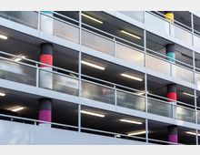 Außenansicht eines mehrstöckigen Parkhauses mit offenen Etagen, sichtbaren weißen Metallgeländern und farbigen Säulen. Die Beleuchtung im Inneren der Etagen ist eingeschaltet, und einige geparkte Autos sind erkennbar.