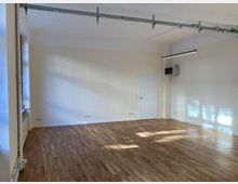 Leerer Innenraum mit hellem Holzboden, cremefarbenen Wänden und einem weißen Türrahmen an der Rückwand. Oben an der Decke verlaufen sichtbare Metallrohre und eine Hängelampe, während durch die Fenster auf der linken Seite Sonnenlicht einfällt.