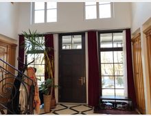 Eingangsbereich eines Hauses mit dunkler Holztür, flankiert von großen Fenstern mit roten Vorhängen. Im Vordergrund steht eine verzierte Metalltreppe, ein Garderobenständer mit Mänteln und einige Topfpflanzen; der Boden ist mit schwarz-weiß karierten Fliesen gestaltet.