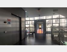 Ein Innenbereich mit einem Aufzug auf der linken Seite, einer Glaswand mit Blick auf ein historisches Gebäude und einem schmalen, mit Geländern versehenen Übergang in der Mitte, der zu einem weiteren Raum führt. Die Umgebung ist modern gestaltet mit dunklen Fliesen und viel Tageslicht.