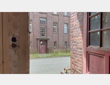 Blick aus einer offenen, verfallenen Tür auf die Fassade eines verlassenen Backsteingebäudes mit großen Fenstern und einer roten Eingangstür; im Vordergrund verwildertes Gras entlang einer asphaltierten Straße.