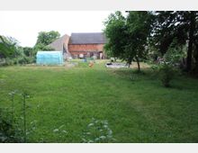 Grüne Wiese mit einzelnen Bäumen, im Hintergrund ein großes Backsteingebäude mit Satteldach. Links steht ein kleines, hellblaues Gewächshaus, daneben Gartengeräte und eine ungenutzte Plane.