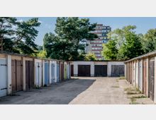 Reihen von flachen Garagen mit verschiedenfarbigen Holztüren auf einem unbefestigten Platz, im Hintergrund Bäume und ein mehrstöckiges Wohngebäude.