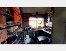 Kleiner, beengter Arbeitsraum mit Regalen an den Wänden, gefüllt mit Werkzeug, Kisten und Geräten. In der Mitte befindet sich ein Schreibtisch mit diversen Gegenständen, darunter eine Kaffeemaschine und Werkzeuge, sowie ein kleines Fenster mit einem halb geschlossenen Vorhang.