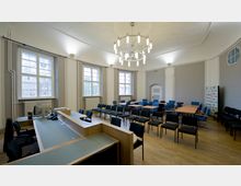 Ein heller Konferenzraum mit hohen Fenstern, Holzboden und weiß gestrichenen Wänden. Der Raum ist mit Tischen, blauen und schwarzen Stühlen sowie einer Empfangstheke ausgestattet; ein Kronleuchter hängt von der Decke.