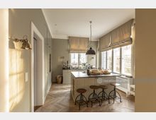 Helle, moderne Küche mit großen Fenstern und viel Tageslicht, einer zentralen Kücheninsel mit vier Barhockern sowie Schränken und Geräten in hellgrauer Farbe. Der Boden ist mit Fischgrätparkett ausgelegt, und an der Decke hängt eine große Metalllampe.