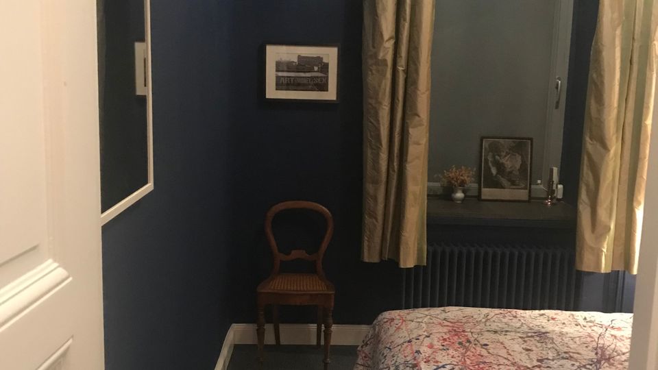 Kleines Schlafzimmer mit dunkelblauen Wänden, einem Bett mit gemusterter Tagesdecke und einem Fenster mit hellen Vorhängen über einem Heizkörper. An der Wand hängen ein Spiegel und ein Bild, daneben steht ein kleiner Holzstuhl; auf der Fensterbank liegen Dekoobjekte.