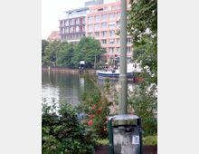 Gebäude mit großen Glasfenstern und roten Fassadenelementen am Ufer eines Flusses, im Vordergrund ein Mülleimer, Büsche mit roten Blumen und ein Laternenmast. Ein Boot ist am Ufer festgemacht, umgeben von dichtem Grün.