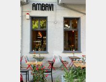 Außenansicht eines Cafés mit der Aufschrift „AMBAVI“ über großen Fenstern, durch die eine gemütliche Inneneinrichtung mit Beleuchtung sichtbar ist. Vor dem Gebäude stehen Holztische mit bunten Stühlen sowie Blumendekoration in einem gepflasterten Außenbereich.