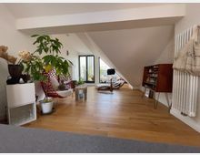 Wohnraum unter einer Dachschräge mit Holzfußboden, großen Fenstern im Hintergrund und moderner Einrichtung. Im Vordergrund stehen Pflanzen, ein roter Stuhl mit Netzdesign, ein Holzregal sowie eine weiße Heizung an der Wand.