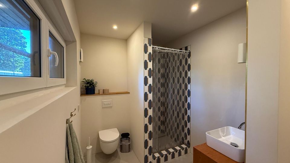 Modernes Badezimmer mit neutralen Beigetönen; ausgestattet mit einer bodenebenen Dusche mit gemusterten Fliesen, einem wandmontierten WC, einem weißen Waschbecken auf einem Holzunterschrank und einem schmalen Fenster auf der linken Seite.