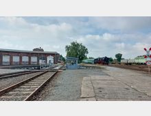 Ein Bahnbetriebswerk mit einem roten Backsteingebäude und mehreren geschlossenen Toren, einer Drehscheibe im Vordergrund und historischen Zügen, darunter eine Dampflok, im Hintergrund. Rechts sind mehrere Gleise mit abgestellten Waggons und ein Bahnübergangsstopp-Schild.