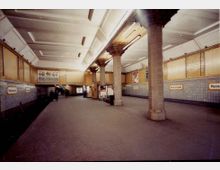 U-Bahn-Station mit rechteckigem Raum, gekennzeichnet durch beige Wände mit Holzverkleidung und darüberliegende blaue Fliesen. Massive, quadratische Säulen und eine abgehängte Decke mit Beleuchtung dominieren die Architektur. Schilder mit dem Stationsnamen „Hermannplatz“ sind an den Wänden angebracht.