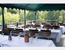 Offene Terrasse mit eingedeckten Tischen, bedeckt von einem grünen Sonnendach. Die Tische sind mit weißen Tischdecken, Kupfergefäßen, kleinen Blumengestecken und umgedrehten Weingläsern dekoriert; im Hintergrund sieht man grüne Bäume.