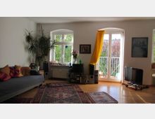 Wohnzimmer mit Holzboden, einer grauen Couch mit bunten Kissen, großen Fenstern und einer Balkontür, die mit einem gelben Vorhang dekoriert ist. Im Raum befinden sich Zimmerpflanzen, Teppiche mit Mustern, ein Fernseher, Bücherregale und eine Stereoanlage.