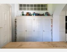 Ein heller Innenraum mit einem großen weißen Einbauschrank in der Mitte, flankiert von offenen Regalen. Oberhalb des Schranks stehen verschiedene Keramiken und Geschirr. Im Vordergrund ist eine helle Holzoberfläche sichtbar, vermutlich ein Tisch.