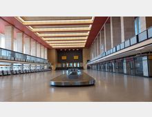 Weitläufige Flughafenhalle mit modernistischer Architektur, geprägt von hohen Säulen, großen Fenstern und einer roten Decke. Zentral steht ein leerer Gepäckförderband, umgeben von Schaltern und Glasfronten.