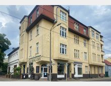 Eckgebäude aus der Gründerzeit mit beiger Fassade und roten Dachziegeln, das im Erdgeschoss Ladenlokale wie „Der Weinladen“ beherbergt. Im Umfeld befinden sich kleinere Gebäude und Bäume, sowie ein Straßenschild und Stromleitungen.