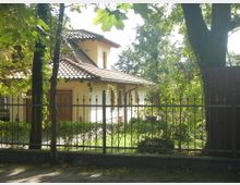 Einfamilienhaus mit hellem Putz und rotem Ziegeldach, umgeben von einem Garten mit Bäumen und einer niedrigen Hecke, eingefasst durch einen schmiedeeisernen Zaun. Im Vordergrund führt ein Gehweg entlang des Zauns.