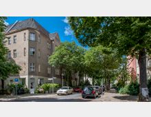 Ecke eines Wohnhauses mit hellbraunem Putz und vielen Fenstern, umgeben von einer baumbestandenen Straße mit parkenden Autos. Der Gehweg ist gesäumt von Bäumen, kleinen Gärten und Schildern, bei klarem Himmel und Sonnenschein.