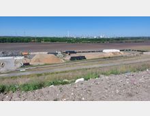 Ein Industriegelände mit Sand- und Schutthaufen, Stapeln von Materialien wie schwarzen und weißen Kunststoffblöcken sowie Maschinen und Fahrzeugen, darunter ein Lastwagen und ein Bagger. Im Hintergrund ist eine weite Ackerfläche und eine Industrielandschaft mit Schornsteinen und Windrädern zu sehen.